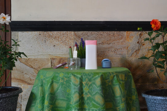 A centered shot featuring a plastic lotion bottle and lipstick on a green fabric tablecloth, framed by flowering plants in the foreground and a small artificial lavender pot behind.