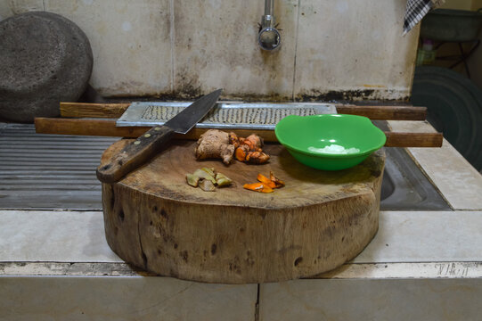 A top-down shot of fresh turmeric and ginger roots being prepared on a wooden cutting board in a kitchen area featuring a metal knife and a traditional grater in the background.