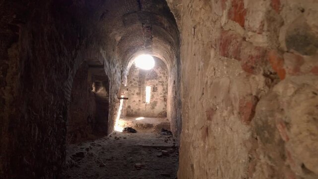 Old underground corridor with rough stone walls and arched ceiling, natural light glowing at end, dusty floor with debris, historic architecture creating moody perspective depth