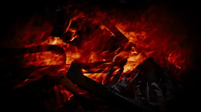 Close up of a burning campfire with bright orange flames and glowing embers on charred wood at night. Intense heat and atmospheric fire light in a dark outdoor environment.