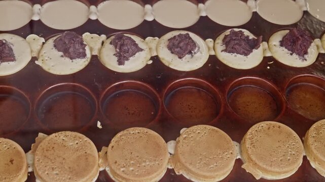 Rows of round Japanese sweet cakes cook in iron molds, some filled with dark red bean paste, others still baking, capturing the traditional street food production process from above.