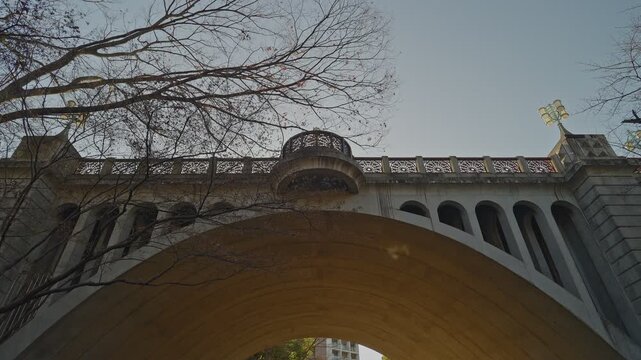 Low-angle static shot through the bridge arch, white ornate lattice parapet and curved balcony spanning above, repeated arch openings right, bare winter branches framing upper left, golden lamp upper