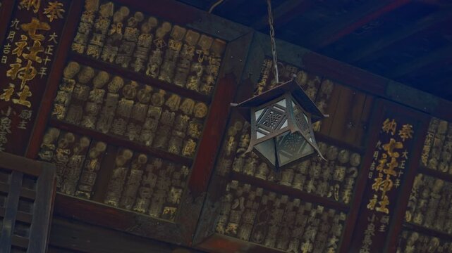 Low-angle upward static shot inside a dimly lit Japanese shrine, ornate hanging lantern centered mid-frame, surrounding walls and ceiling densely covered in aged wooden votive carvings and gilded kanj