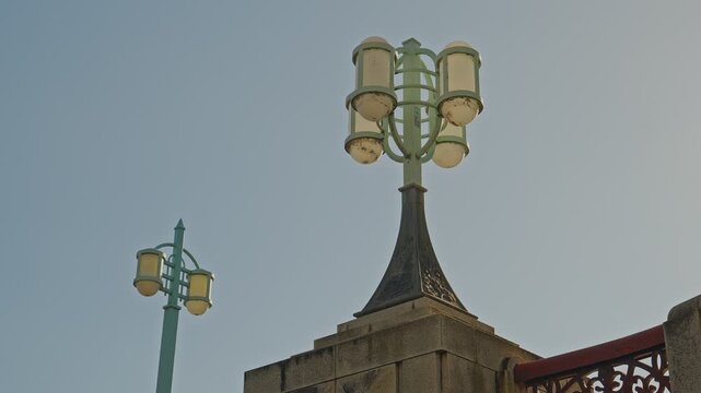 Wide static shot of two ornate vintage multi-globe lamp posts atop a historic stone bridge parapet, green metal stems, decorative pillar finial in foreground, pale grey overcast sky behind.