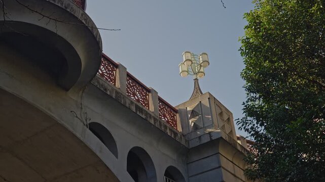 Low-angle static shot of a historic stone bridge parapet, red lattice railing curving left, elegant multi-globe lamp post centered against clear blue sky, green tree foliage framing the right edge.