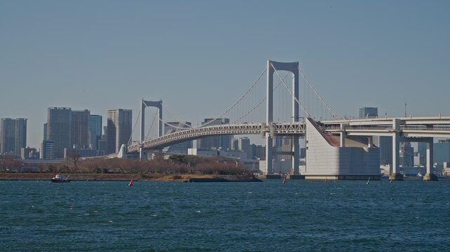 Rainbow Bridge's arch span and suspension cables fill the frame over Tokyo Bay, with a barge, wooded island, and dense city skyline behind, captured as a static medium wide shot in clear daylight.