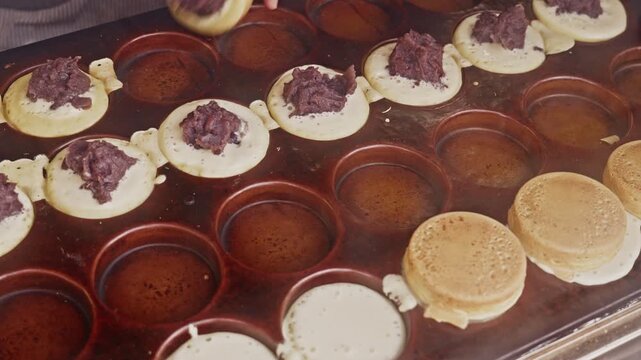 A vendor's hand places a baked cake half onto red bean paste-filled rounds on a hot iron griddle, capturing the hands-on assembly process of traditional Japanese imagawayaki street food.