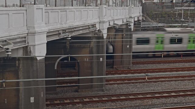 A green and white commuter train appears from behind aged brick arch bridge structures along a wide multi-track railway yard. Overhead wires and layered rail infrastructure reinforce the dense, histor