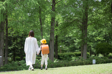 Fototapeta premium 新緑の公園を手をつないで歩く母親とランドセル姿の女の子 親子の登校風景全身広角