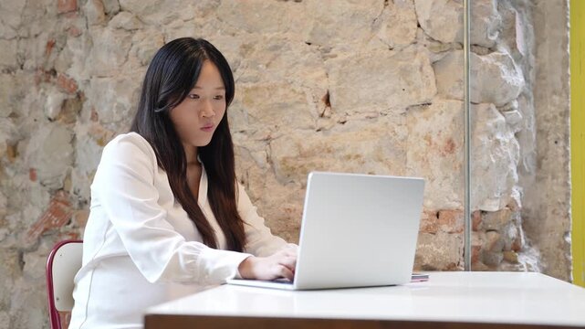 Young woman smiles while typing on laptop. Her hands move confidently across the keyboard. She sits at a simple desk near a stone wall. Natural light streams through the window
