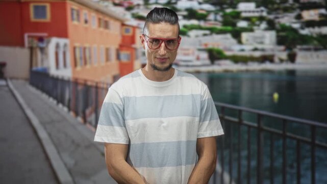 Man with red glasses and goatee makes finger to lips shushing gesture on a waterfront street beside metal railing; quiet curiosity.