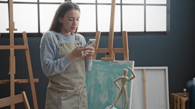 Woman painter in apron holds smartphone with both hands tapping screen beside turquoise canvas and wooden mannequin in studio; focused.