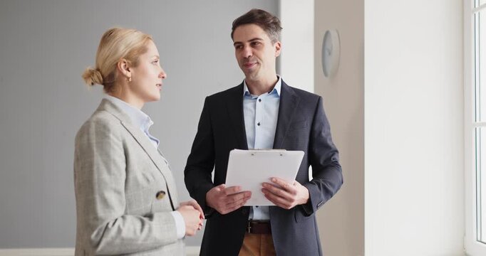 Realtor and client in apartment viewing. Agent consults by window holding clipboard, explaining features, pricing and timelines for housing decisions. Concept conveys trusted real estate.
