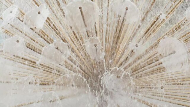 Water fountain detail showing dandelion shape spraying droplets in sunlight with motion blur effect of splashing streams creates refreshing urban atmosphere during summer day in public city square.
