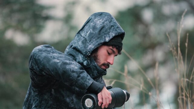 A man in a hooded jacket drinks mate from a gourd with a metal straw, surrounded by winter foliage. Ideal for themes of outdoor adventure and winter camping.