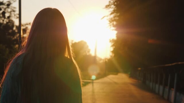 Woman walking on country road during beautiful sunset, slow motion footage of young girl strolling outdoors with long hair fluttering in wind, golden hour light illuminating path.