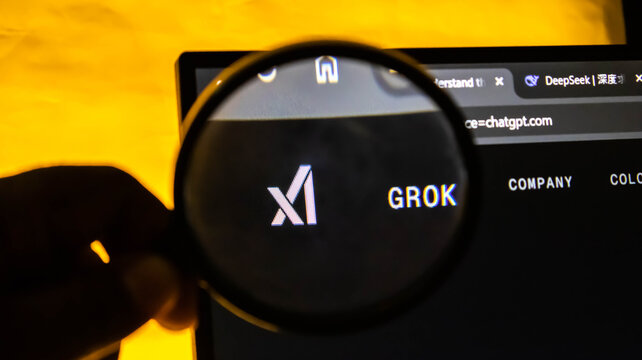 Closeup of a magnifying glass held over a computer screen displaying the xai grok logo and website interface with a warm yellow background lighting