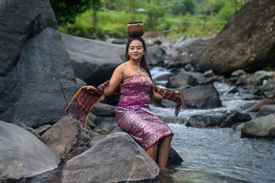 Elegant Asian woman in batik dress dancing pose with clay pot on head near tropical stream