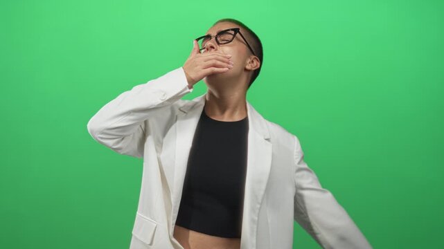 Woman with midriff exposed covering mouth with hand in studio wearing glasses, white blazer and black crop top against green backdrop; tiredness solitude.