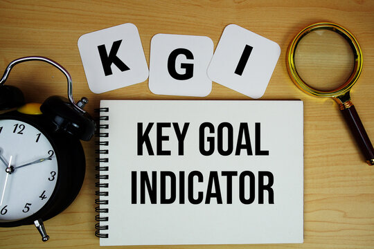 KGI Key Goal Indicator text written on notepad with magnifying glass and alarm clock on wooden background