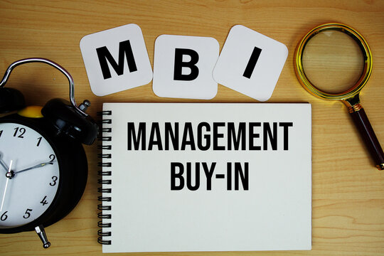 MBI Management Buy-In text written on notepad with magnifying glass and alarm clock on wooden background