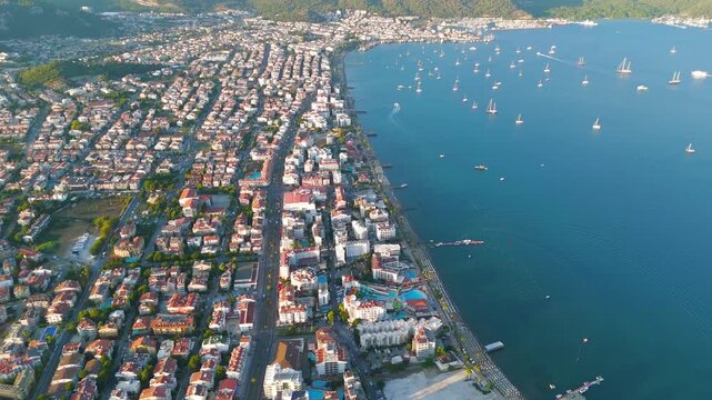 Marmaris, Turkey. Aerial view of dense coastal resort grid with red-tiled roofs stretching along a yacht-dotted bay, backed by pine-clad mountains at golden hour. Aerial View