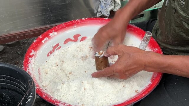 An Asian man selling traditional food fills a bamboo mold with Putu cake batter made from rice flour and palm sugar before the steaming process begins.