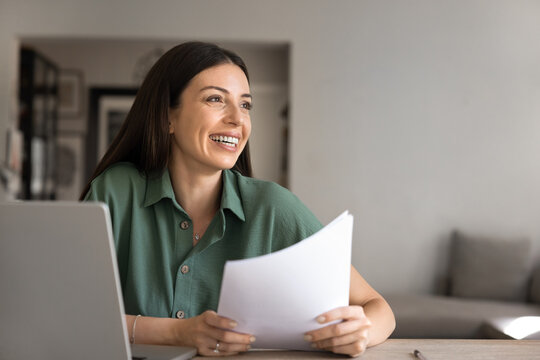 Woman holds papers documents at home office desk, distracted from information review, smiling, enjoy good news, positive notification, feels satisfaction with remote work result, achievement, success