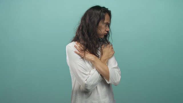 Woman hugging shoulders with crossed arms in studio wearing white blouse, tense expression and closed posture conveying self comfort; anxiety vulnerability.