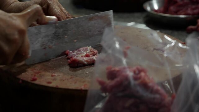 Vendor finely slices raw meat with a sharp knife on a round wooden chopping block while preparing small pieces of meat for cooking. Precise slicing.