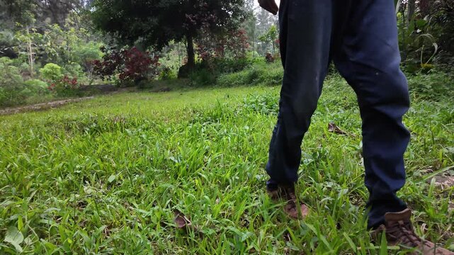 A side-view close-up of feet wearing brown hiking boots and blue trousers walking through a lush green grass field.