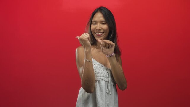 Young thai woman smiles holding bent fingers in a playful paw gesture, showing bare arms and bracelets against a red studio wall; playful joy.