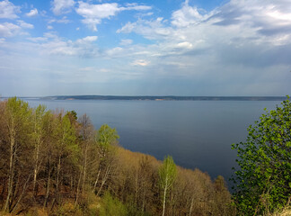 Obraz premium A scenic view of a calm lake bordered by spring trees, under a partly cloudy sky with soft sunlight