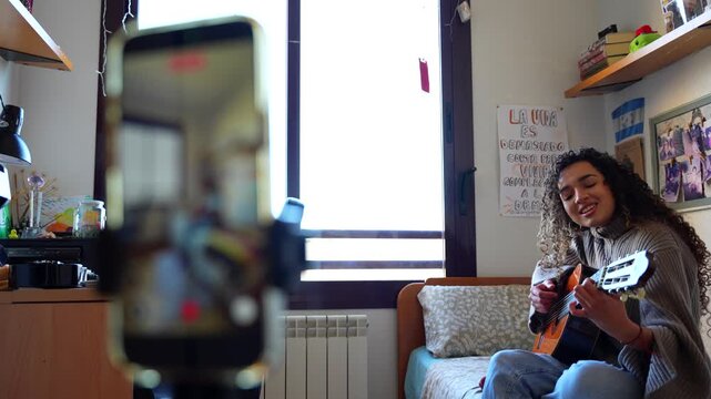 Creative female musician singing and playing guitar while vlogging in her bedroom for social media