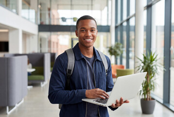 Fototapeta premium Portrait of a happy male university student holding a laptop on a modern college campus