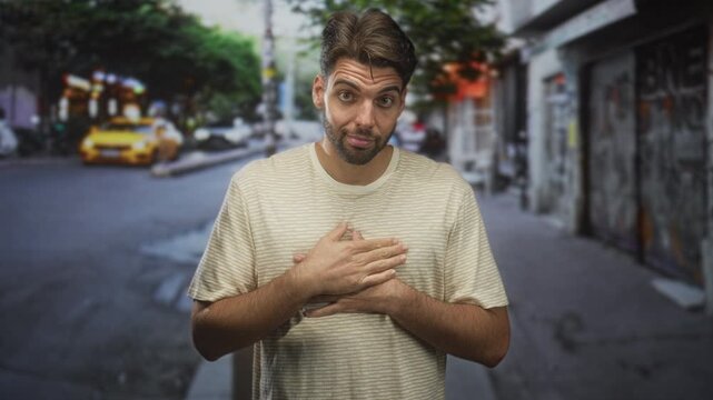 Hispanic man with beard placing hands on chest in a graffiti lined urban street, taxi passing; apology humility.