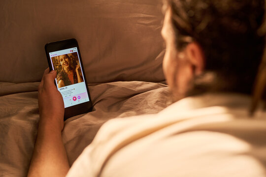 Over shoulder view of young adult man browsing dating app on smartphone in bed, swiping profile match. Concepting online romance, useful for digital dating or relationship marketing