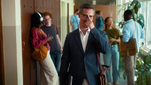 Portrait of a Confident male professor with glasses smiling at camera in university hallway with diverse students in background during class break.