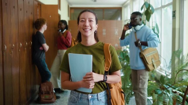 Portrait of a Smiling Korean female student with backpack and notebook looking at camera in university hallway during break with diverse classmates in background.