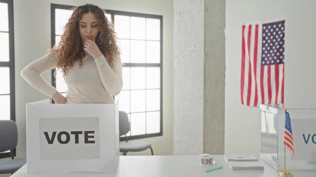 Woman inserts ballot into vote box at polling table in building, leaning over the privacy booth with registration forms and pen visible; thoughtful civic duty.