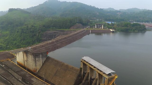 Editorial aerial screenshot showing Saguling Dam concrete wall, floodgate spillway, maintenance roadway, reservoir shoreline and hydroelectric office facility May 2026