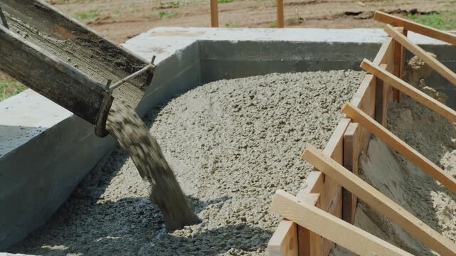 Concrete pouring into wooden formwork on construction site for building foundation during bright daylight shows liquid cement mixture flowing from chute creating solid structure for house development.