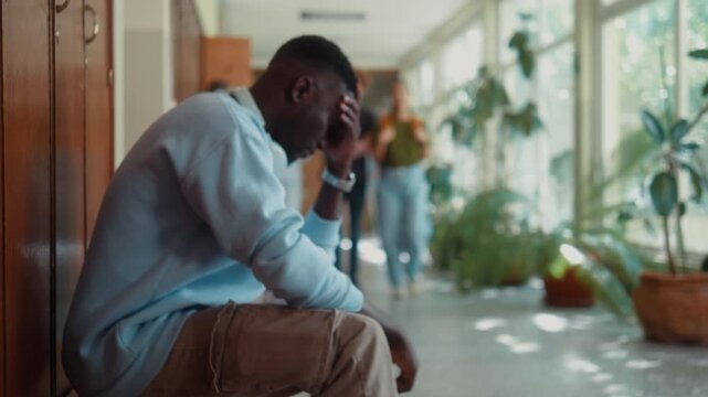 Upset African university student throws backpack on bench after failing exam while supportive Asian classmate comforts him in campus hallway during break.
