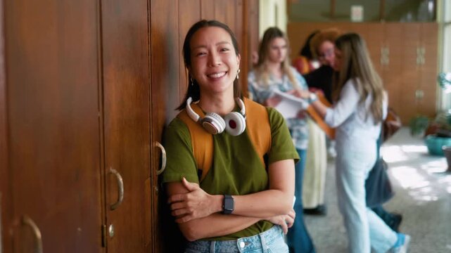 Smiling Korean female student leaning on lockers in university hallway during break and loking at camera with smile. Happy campus lifestyle portrait with diverse students in background.