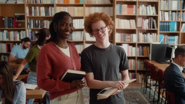 Portrait of smiling Caucasian male student and Black female student holding books in university library while looking at camera.