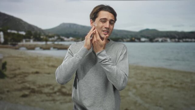 Man touches cheek and jaw with hands, wincing while checking skin by a seaside building; irritation grooming routine.