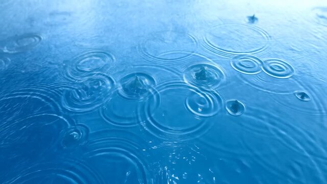 Slow motion top down static shot of raindrops creating ripples on blue water surface in pool outdoors during overcast morning
