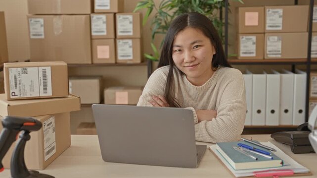 Woman smiling at laptop with folded arms inside a small business building, packages and shelves visible, barcode scanner on desk; small business confidence.