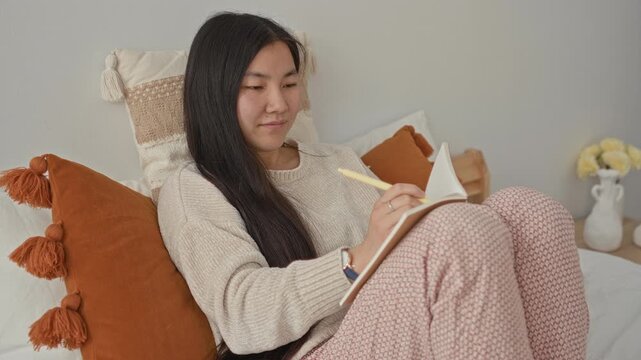 Woman writing in notebook on bed, holding pen and book while reclining among pillows in a building; calm reflection.