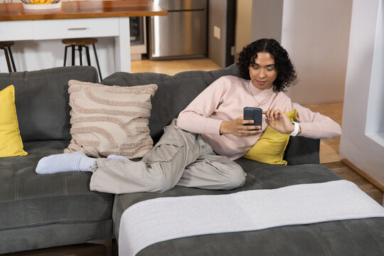 Non-binary adult reclining on gray sectional sofa near kitchen counter, scrolling smartphone